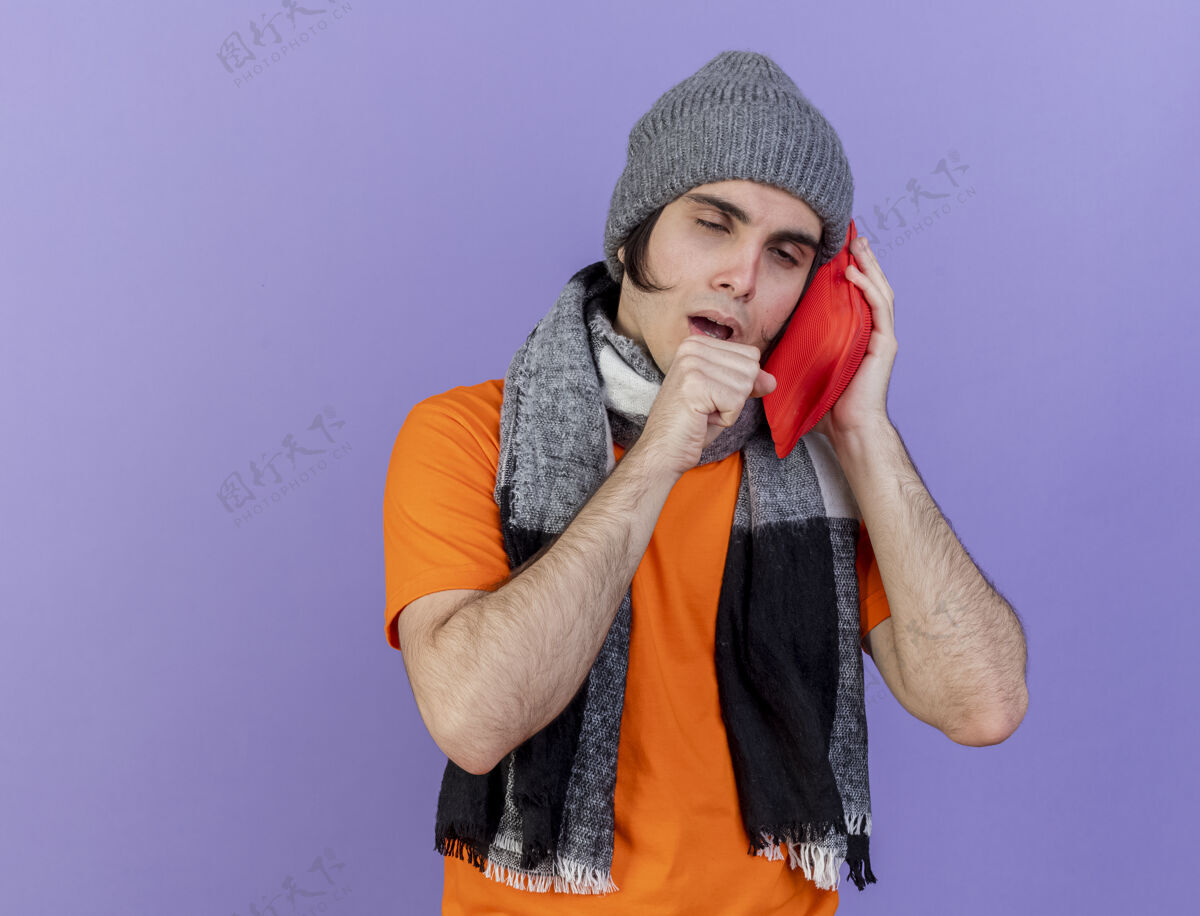帽子咳嗽的年轻病人戴着冬天的帽子 戴着围巾 把热水袋放在脸颊上 隔离在紫色的背景上咳嗽年轻包