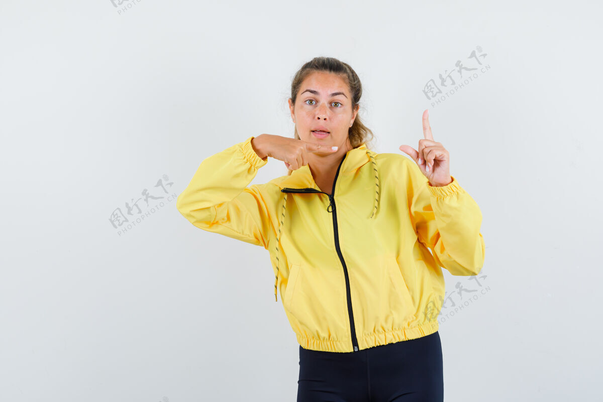 黑发金发女人举着食指做着尤里卡手势 穿着黄色棒球服和黑色裤子指着它 看起来很开心人肖像前面