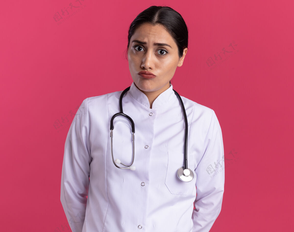 认真的身穿医用外套的年轻女医生带着听诊器站在粉红色的墙上 严肃地看着前方脸听诊器年轻人