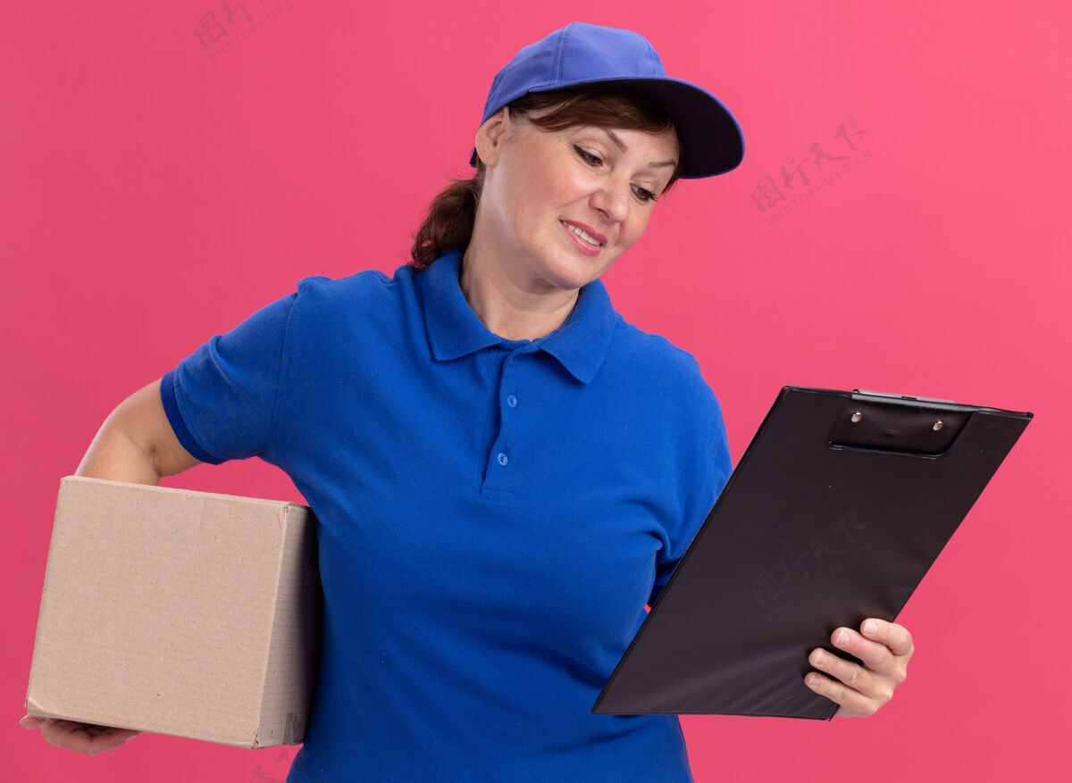 拿着身穿蓝色制服 头戴鸭舌帽的中年女送货员拿着纸板箱和剪贴板站在粉色的墙上微笑着看着它送货剪贴板站着