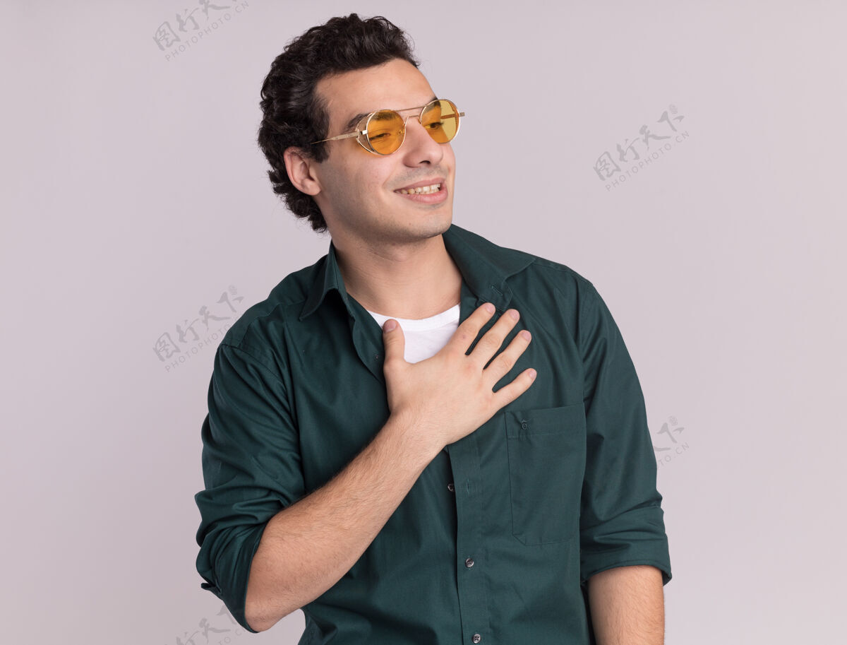 胸脯一个身穿绿衬衫戴眼镜的年轻人站在白色的墙上 一边看着一边 手放在胸前 微笑着 心怀感激感觉眼镜放在一边