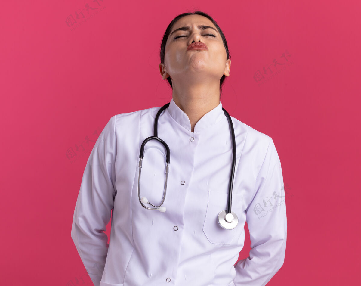 医生穿着医用外套的年轻女医生 带着听诊器 闭着眼睛 站在粉红色的墙上 脸上露出恼怒的表情站立外套眼睛