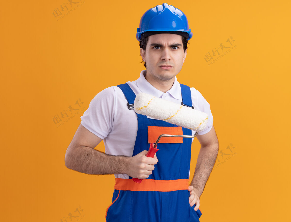 男人年轻的建筑工人穿着建筑制服 戴着安全帽 拿着油漆辊 严肃的脸站在橙色的墙上 看着前面建筑工人站着抱着