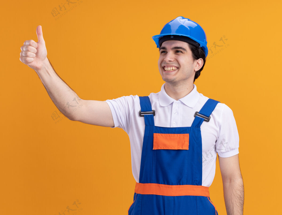 安全年轻的建筑工人身穿建筑制服 戴着安全帽 面带微笑 站在橙色的墙上竖起大拇指拇指建筑微笑
