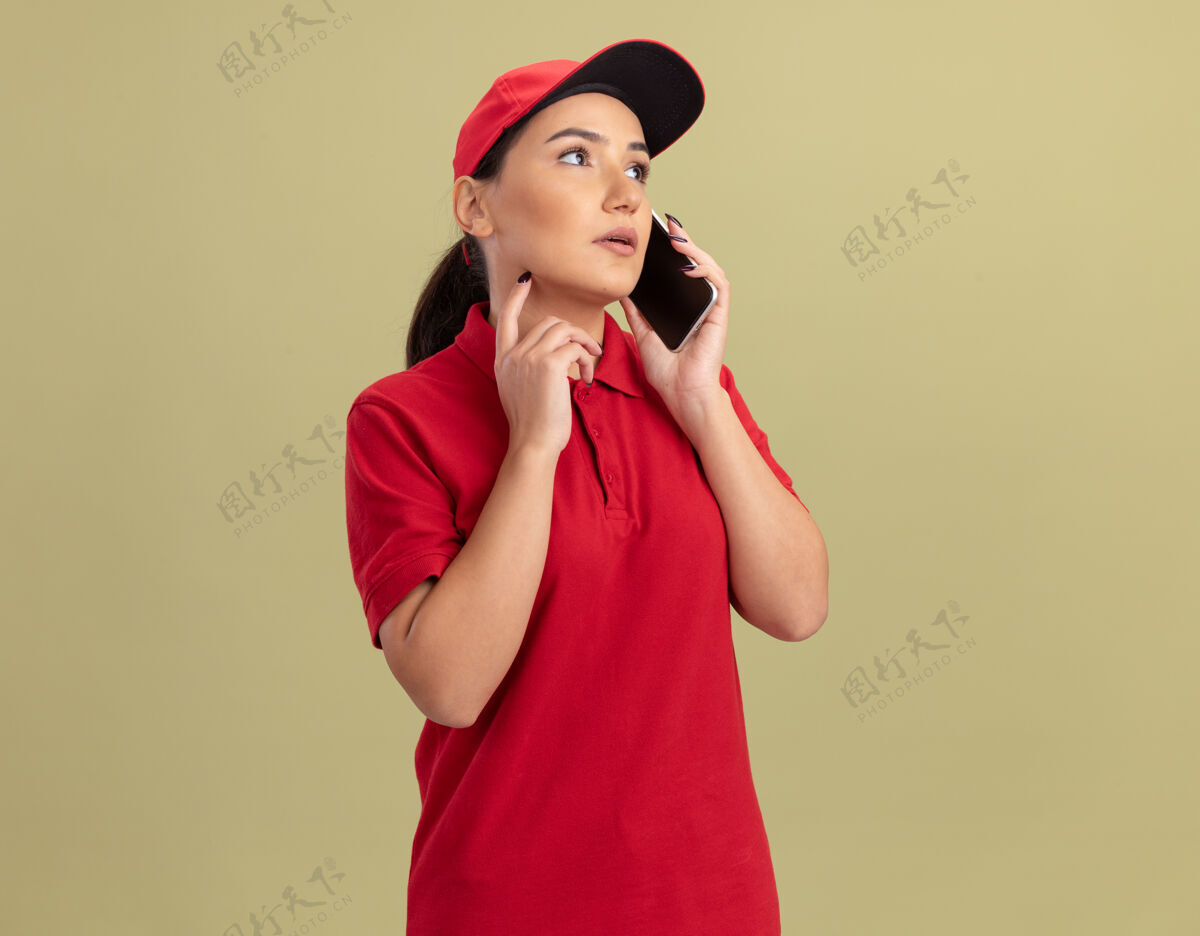 送货身穿红色制服 戴着帽子的年轻送货员站在绿色的墙上 严肃地讲着手机电话严肃制服