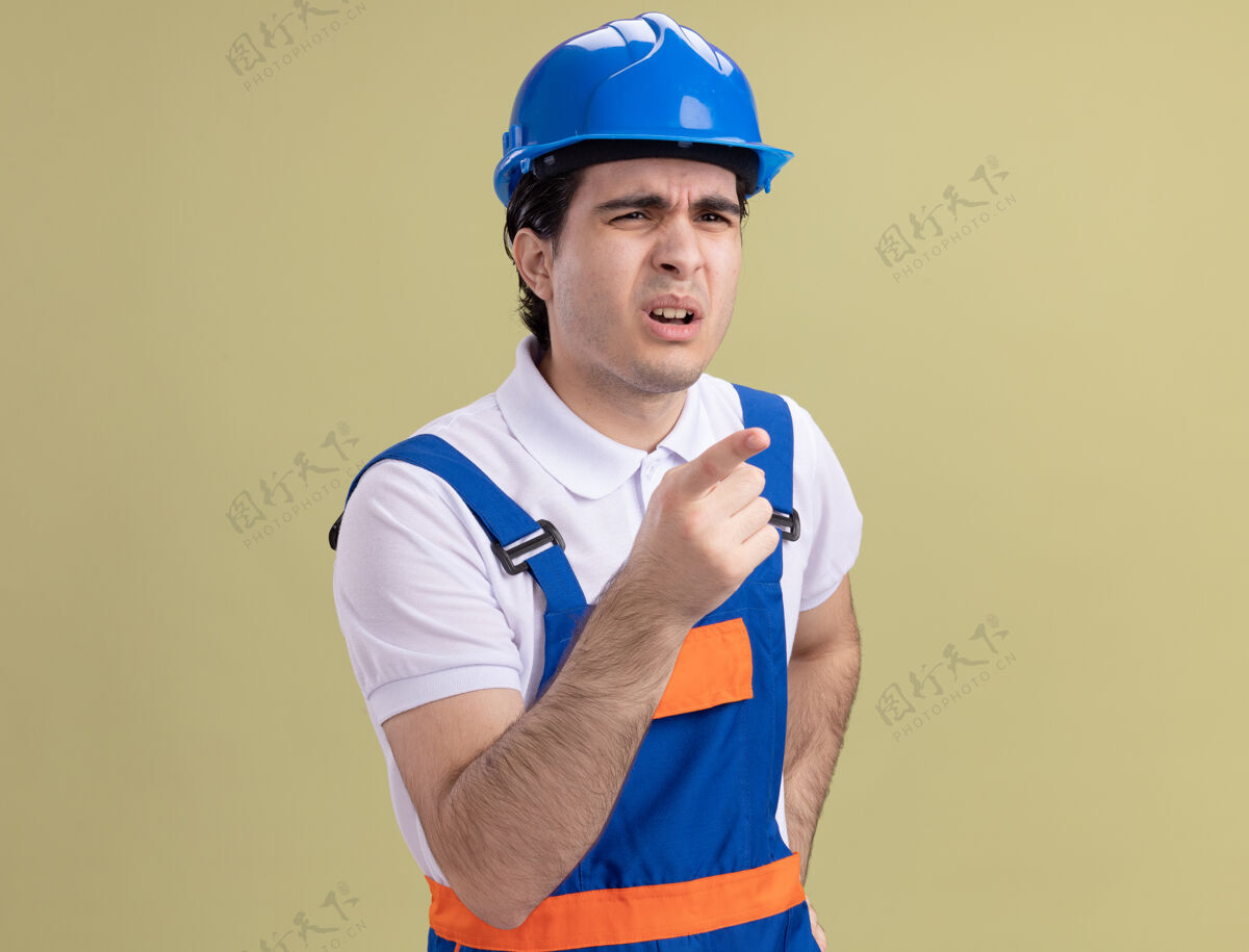 制服年轻的建筑工人穿着建筑制服 戴着安全帽 表情混乱地看着旁边 用食指指着绿色墙壁上的东西头盔困惑某物