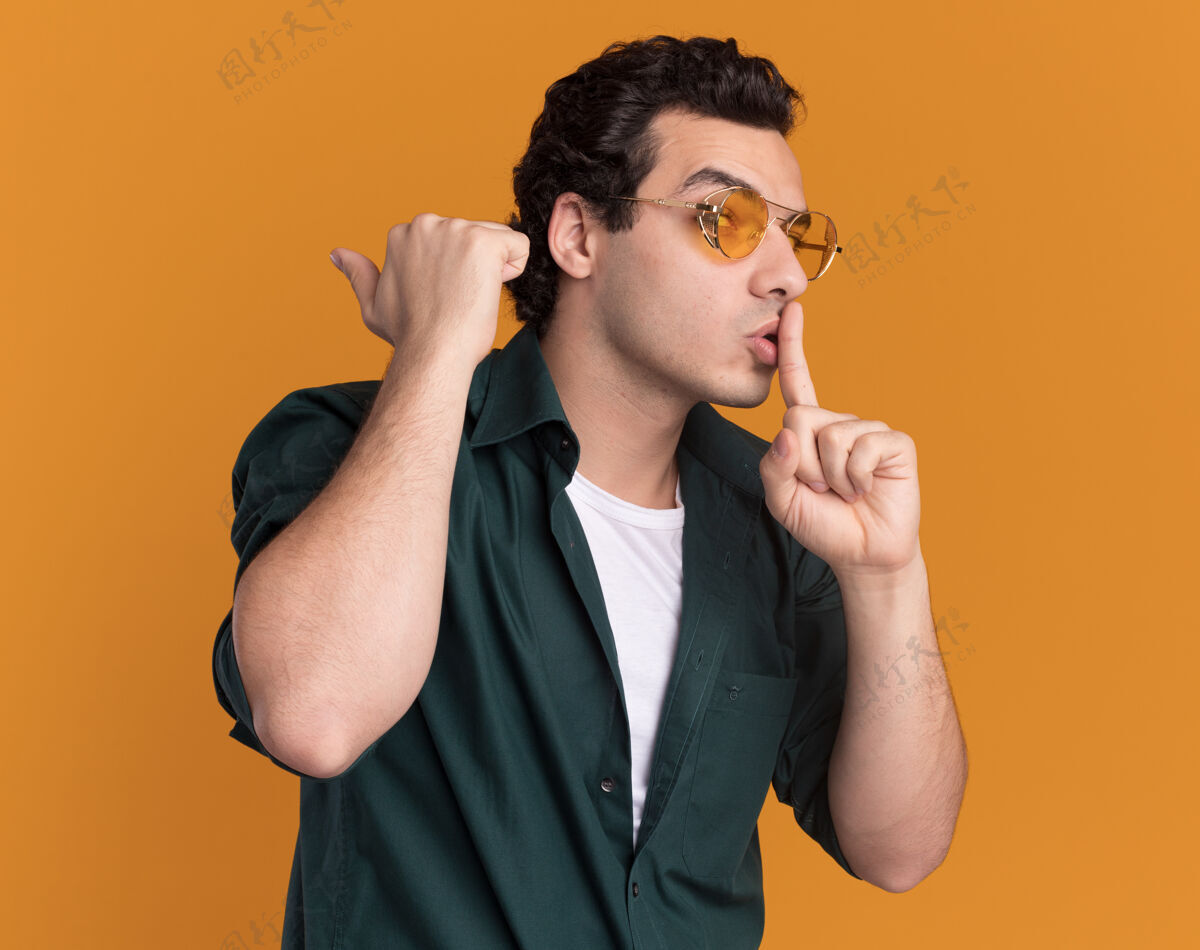 做着身穿绿色衬衫 戴着眼镜的年轻人站在橙色的墙上 手指放在嘴唇上做着沉默的手势沉默眼镜放在一边