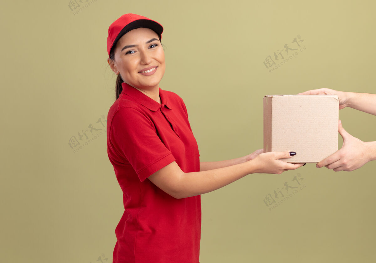 制服身着红色制服 头戴鸭舌帽的年轻女送货员站在绿色墙壁上 微笑着向顾客赠送纸板箱帽子女人送货