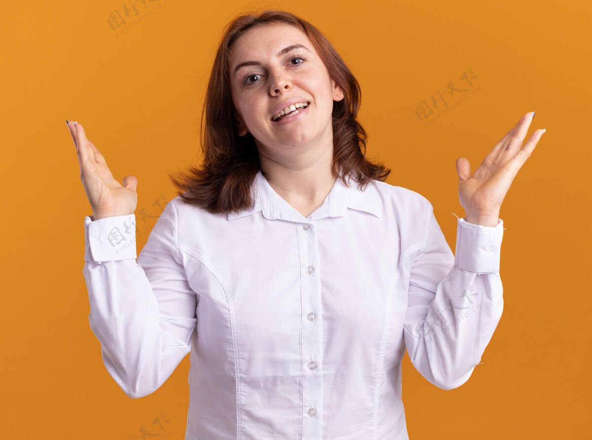 女人身穿白衬衫的年轻女子站在橘色的墙上 面带微笑 高举双臂看着前方衬衫欢呼举起