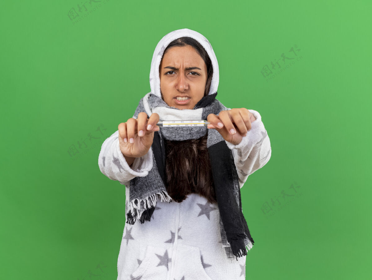 女孩忧心忡忡的小女孩戴着头巾 戴着围巾 对着隔离在绿色背景上的摄像机 拿着温度计相机关心疾病