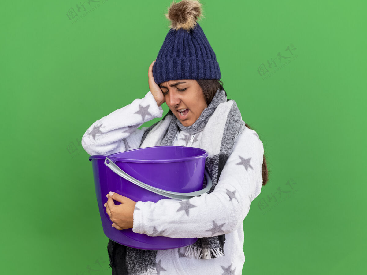 不愉快戴着冬天的帽子戴着围巾拿着塑料桶手放在眼睛上的未被释放的生病的年轻女孩在绿色背景上孤立年轻持有水桶