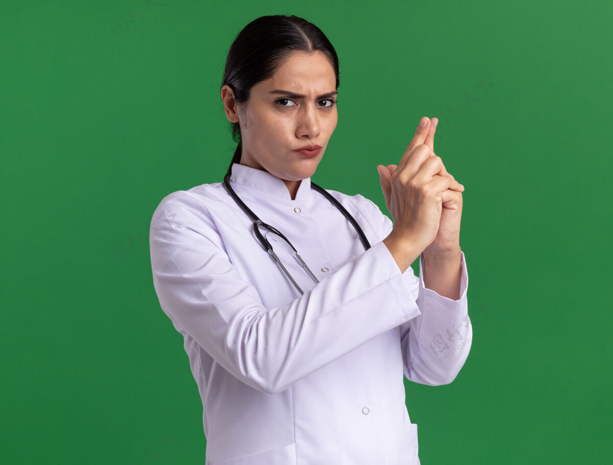 女人年轻的女医生穿着医用外套 拿着听诊器 严肃地看着前面 用手指站在绿色的墙上做着一个手枪手势听诊器做医生
