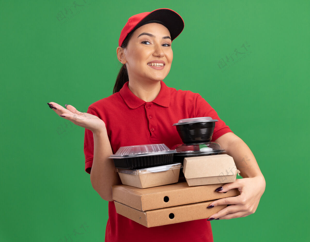 递送身穿红色制服 头戴鸭舌帽的年轻女送货员举着比萨饼盒和食品包 面带微笑地看着前面 手臂举过绿色的墙壁盒子脸站着