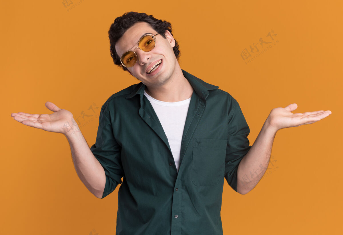 男人身穿绿色衬衫 戴着眼镜的年轻人面带微笑地站在橙色的墙上 向两边伸出双臂站着眼镜穿上