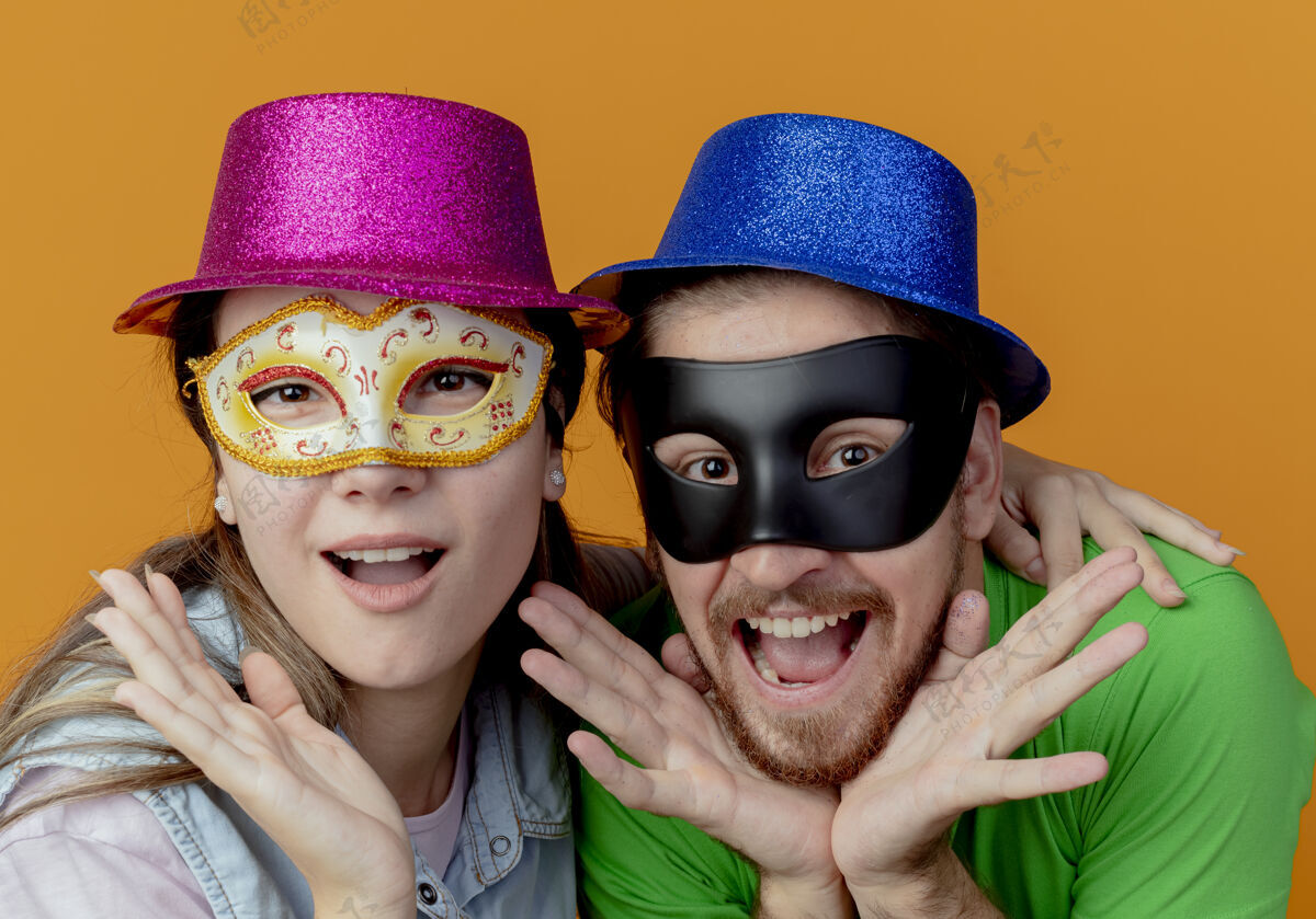 狂欢节年轻兴奋的夫妇戴着粉色和蓝色的帽子戴上化装眼罩把手放在隔离在橙色墙上的下巴上橙色派对面具