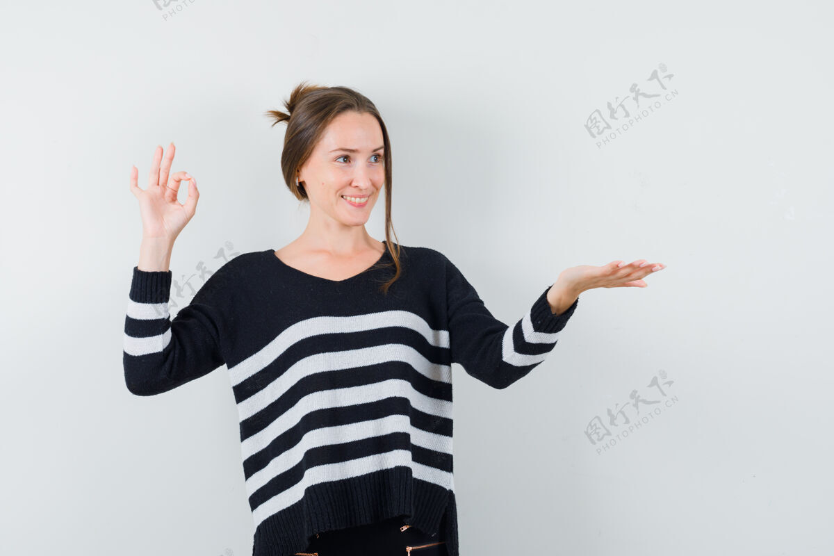 年轻一个年轻的女人展示着ok标志 伸出一只手拿着一件条纹针织衫和黑色裤子 看起来很开心东西美丽漂亮