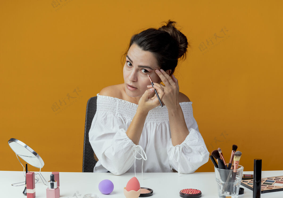 化妆品惊艳的美女坐在桌旁 用化妆工具用眼影和化妆刷隔开橙色墙惊喜眼影桌子