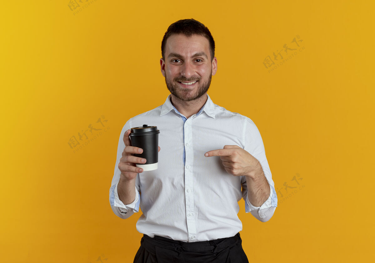 点笑容可掬的帅哥拿着咖啡杯指着隔离在橙色墙上的咖啡杯咖啡年轻杯子