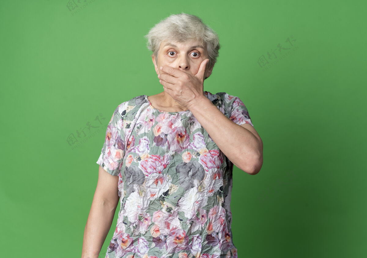 老震惊的老妇人把手放在嘴上 看起来孤立无援地站在绿色的墙上老老手