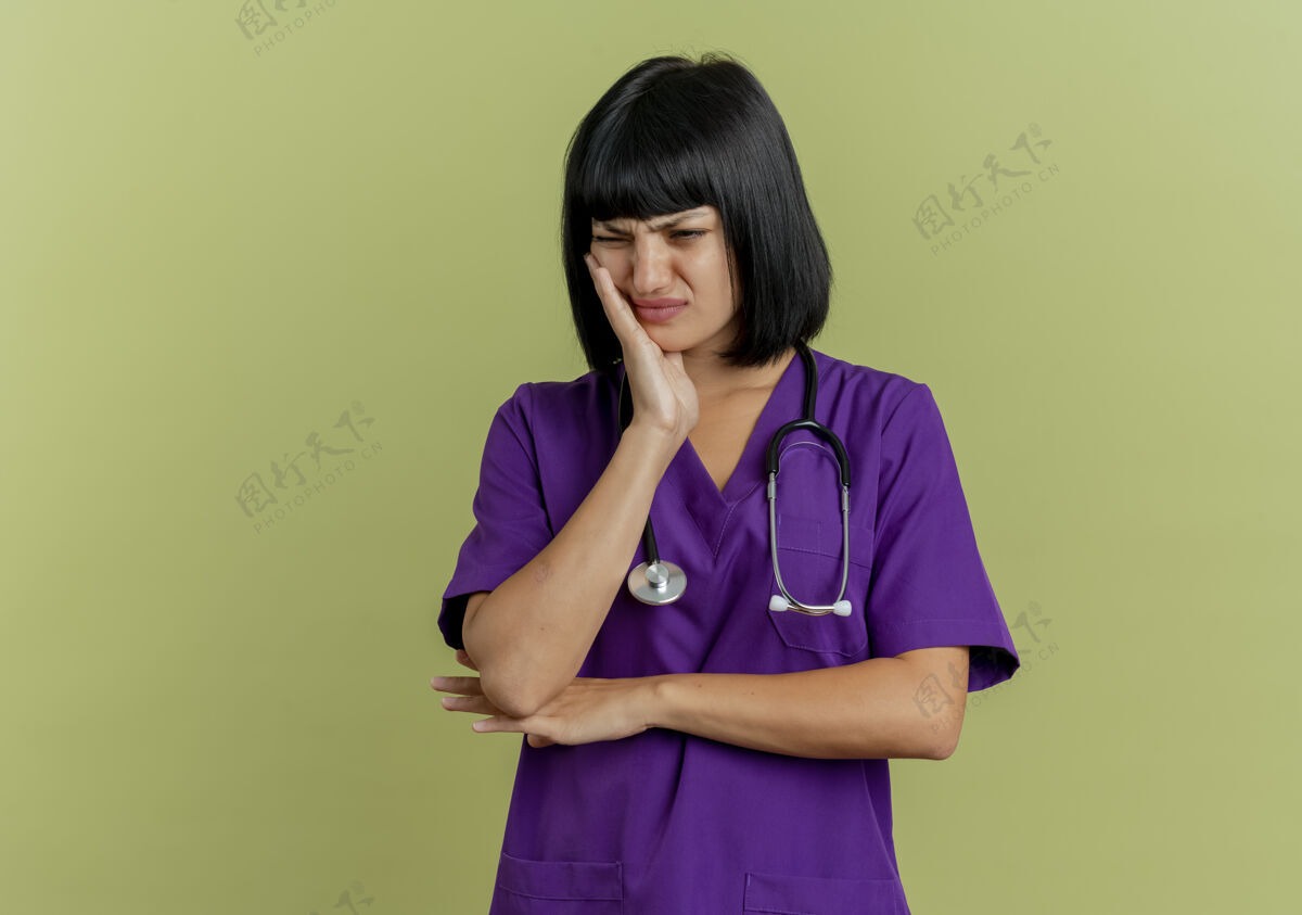 听诊器疼痛的年轻黑发女医生穿着制服 用听诊器将手放在脸上 隔离在橄榄绿的背景和复印空间上年轻黑发女人