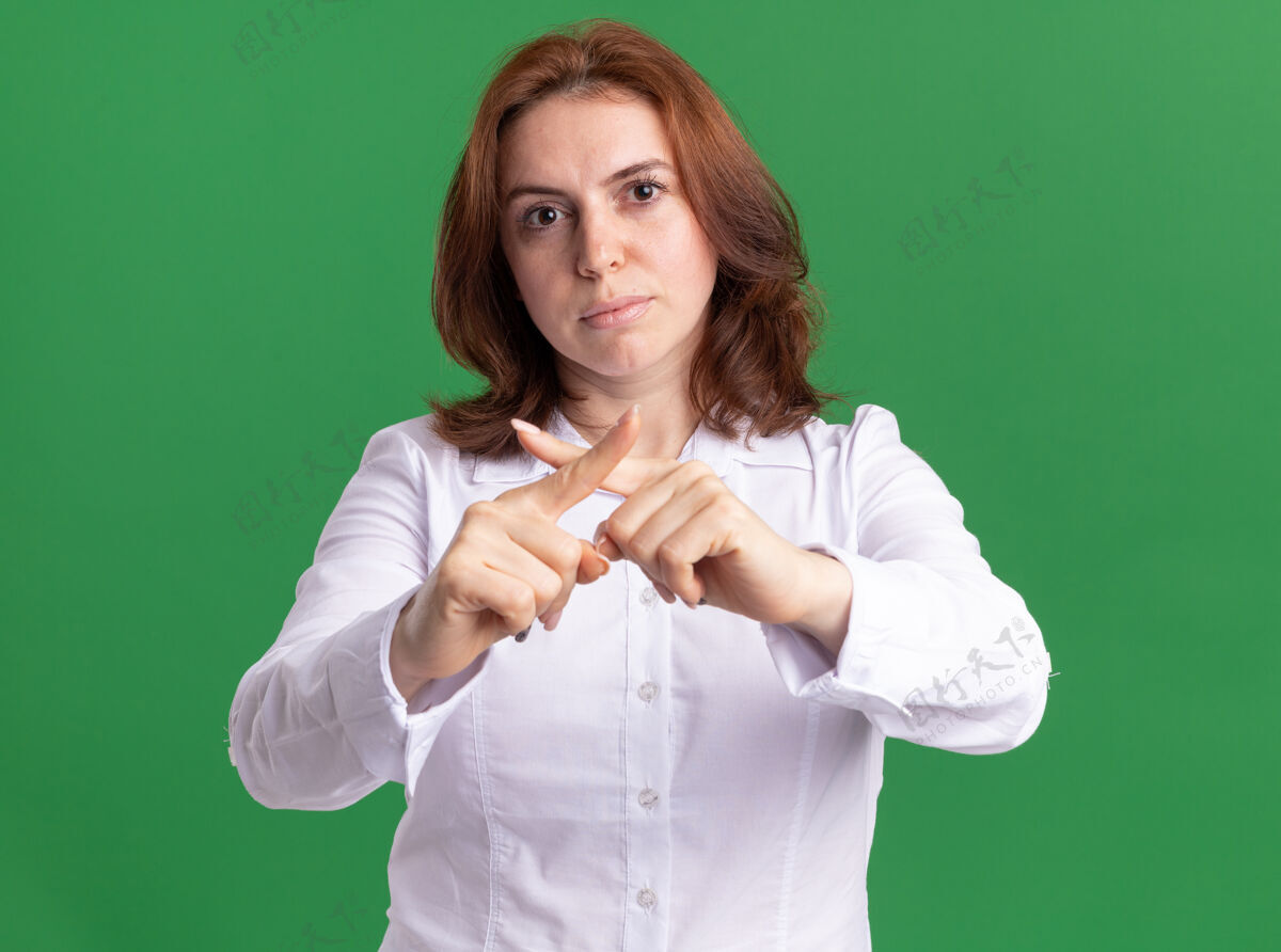 女人穿白衬衫的年轻女子严肃地看着前面 停止手势 交叉食指站在绿色的墙上脸年轻目录