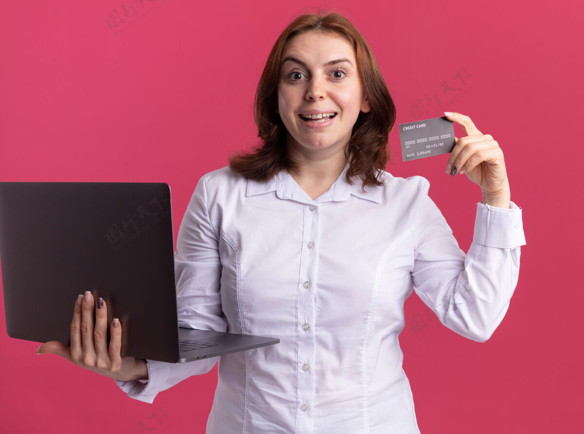 年轻穿着白衬衫的年轻女子拿着笔记本电脑 看着前面的信用卡 站在粉色的墙上 她笑得很开心信用微笑笔记本电脑