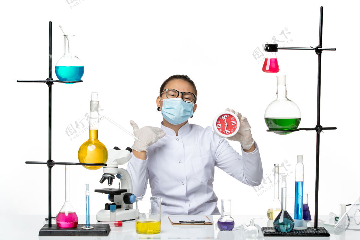飞溅前视图穿着医疗服的女化学家 戴着面具 白色背景上拿着红色的时钟 飞溅着病毒化学的病毒-实验室套装前面时钟