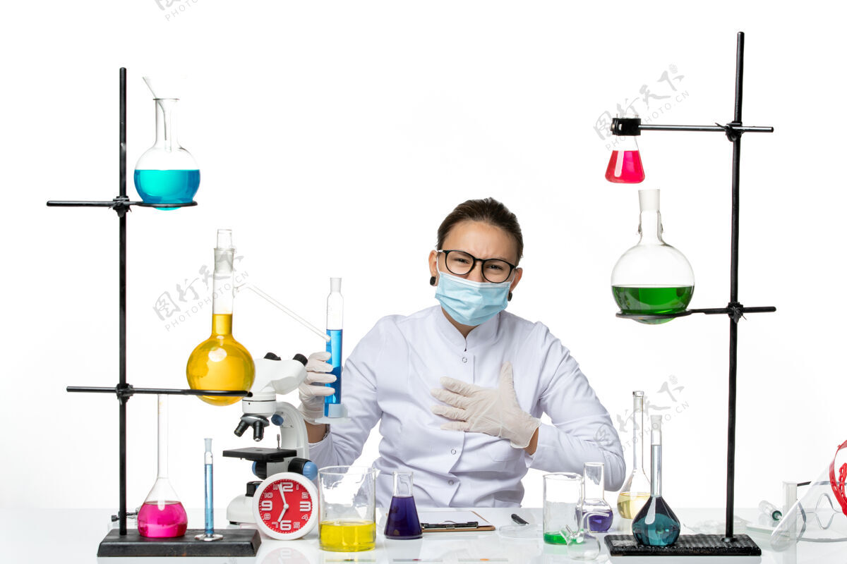 烧瓶前视图穿着医疗服的女化学家 戴着面罩 拿着白色背景上蓝色溶液的烧瓶 溅起化学实验室的水花医学医生解决方案