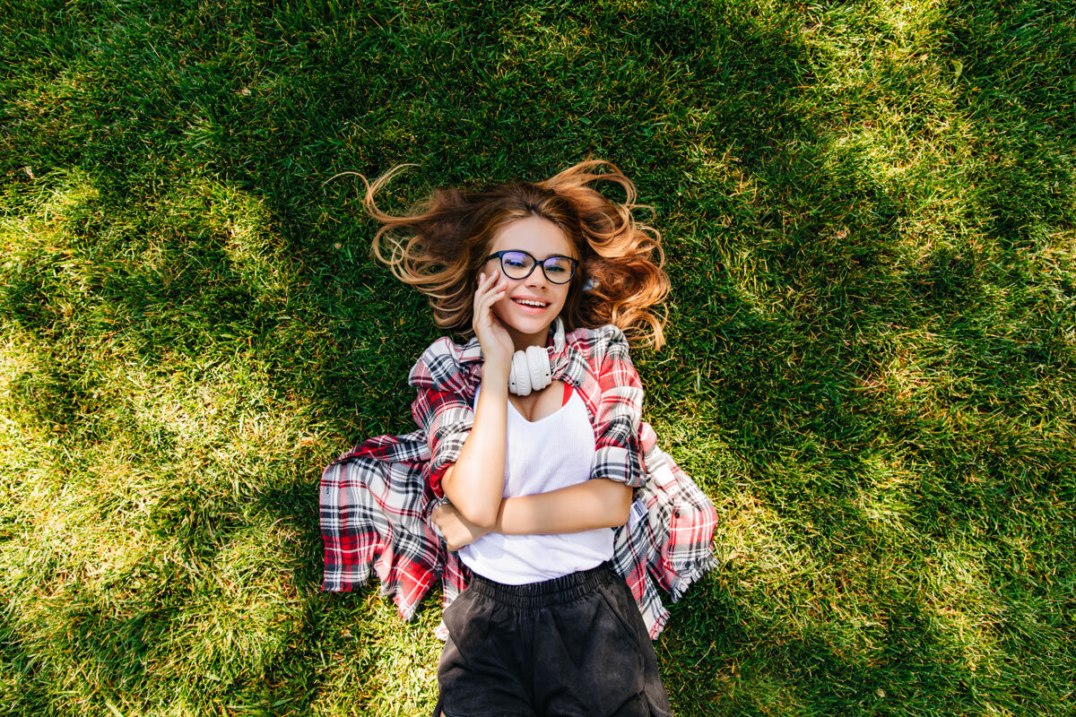 金发美丽的高加索女孩戴着眼镜躺在绿色的草坪上头顶上的户外肖像是一个可爱的年轻女子在公园里发冷头顶欢乐公园