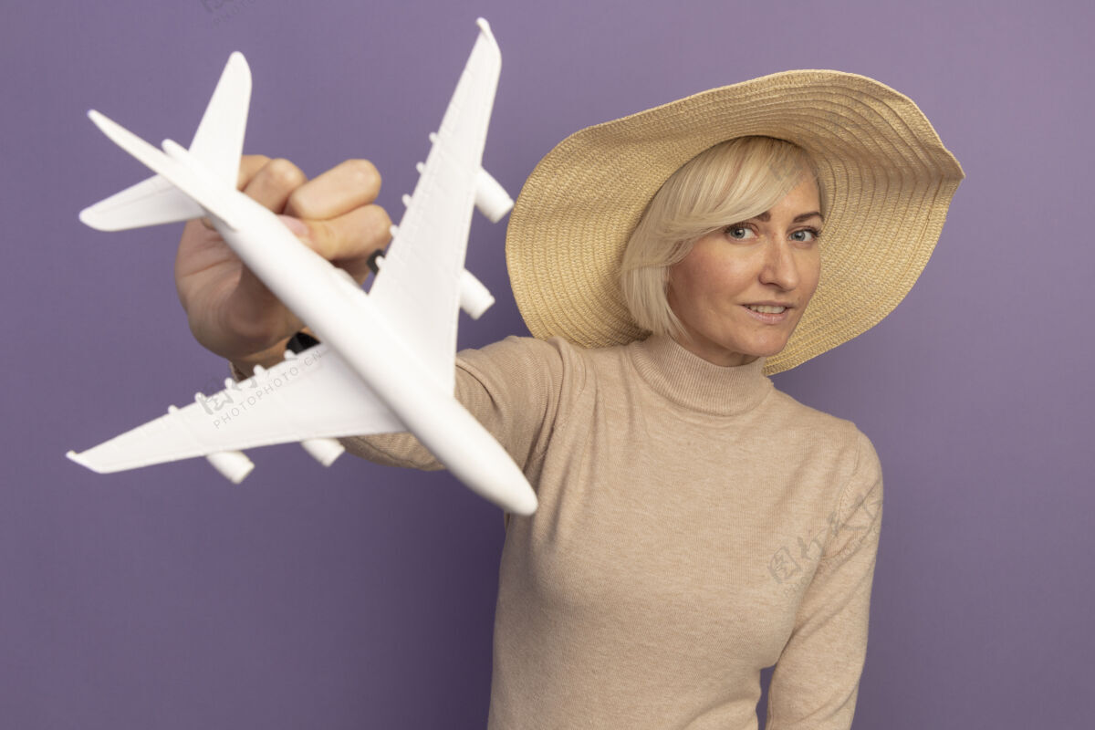请戴着沙滩帽的漂亮金发斯拉夫女人拿着紫色的飞机模型时尚帽子时尚
