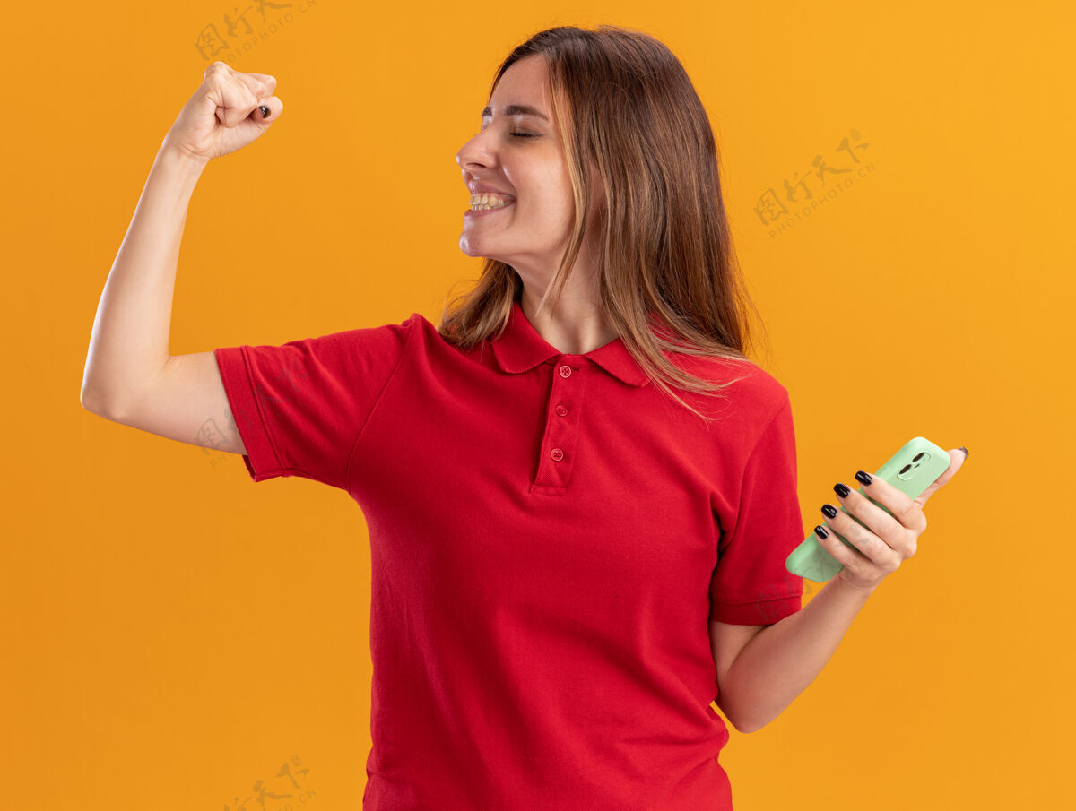 成年人年轻漂亮的白人女孩微笑着握紧拳头 拿着橙色的手机漂亮电话人