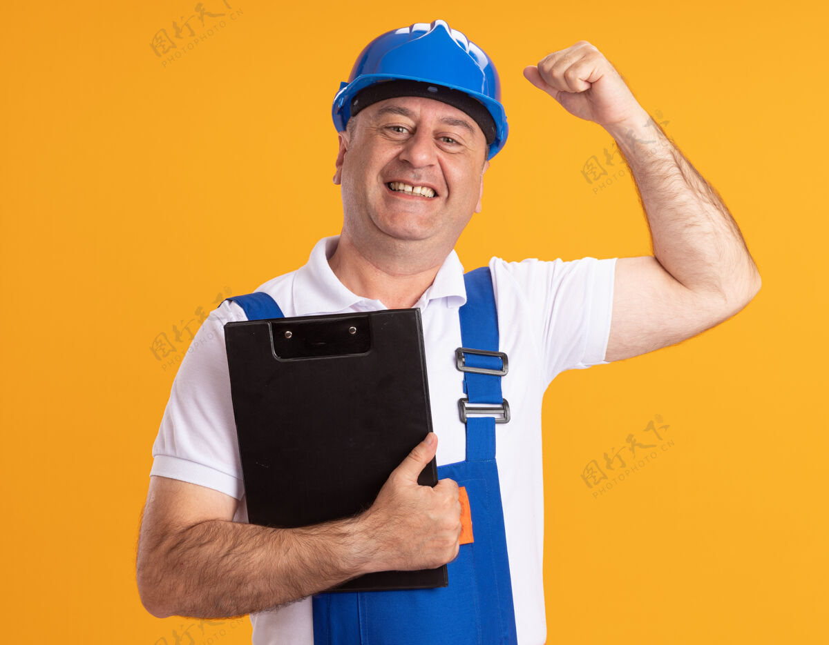 男人身着制服的高加索成年建设者微笑着拿着剪贴板 举起拳头对着橘子人拳头制服