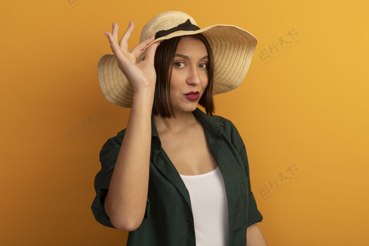沙滩自信漂亮的高加索女人戴着沙滩帽把手放在橙色的帽子上漂亮帽子姿势