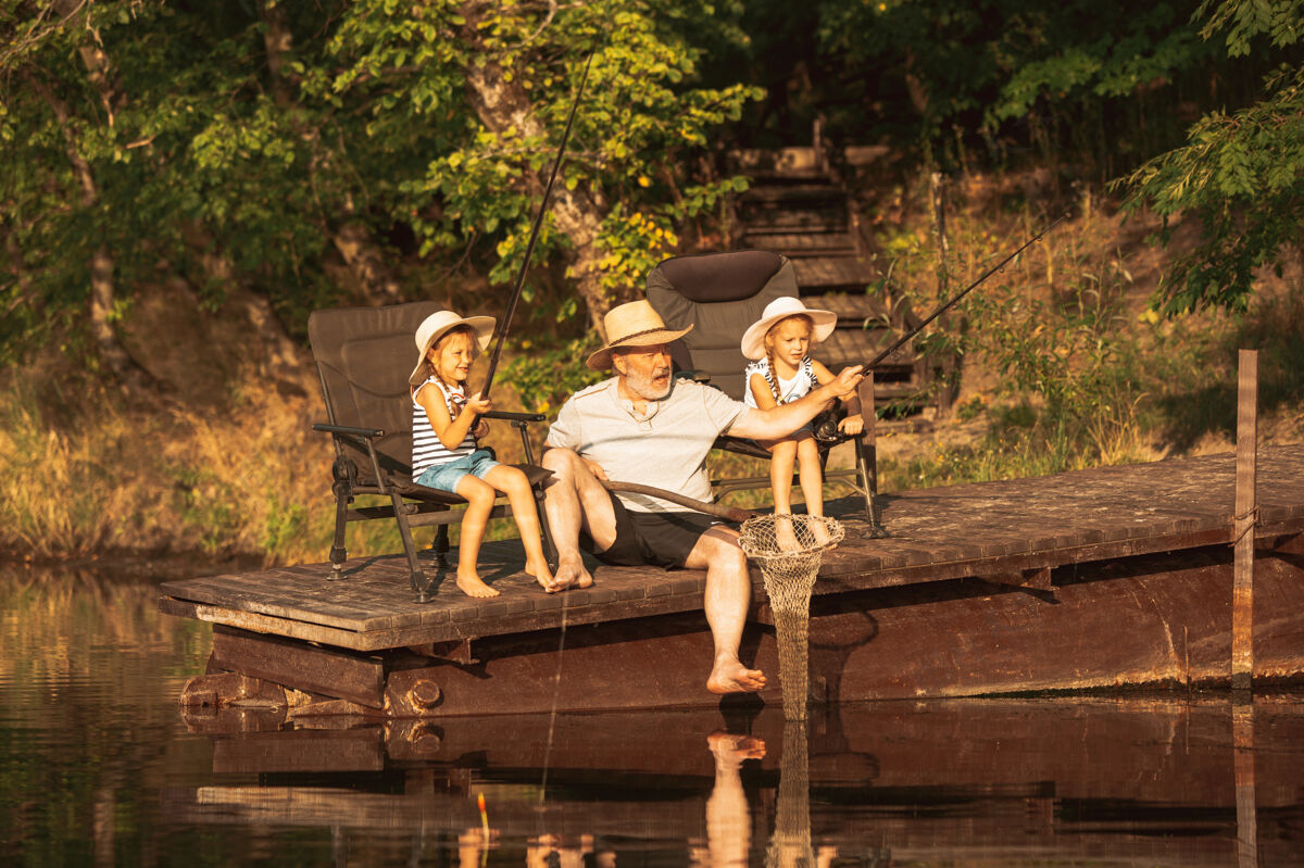 夏天可爱的小女孩和他们的爷爷在湖边或河边钓鱼在夏天的日落时分 在靠近水和森林的码头上休息家庭 娱乐 童年 自然的概念自然玩鱼
