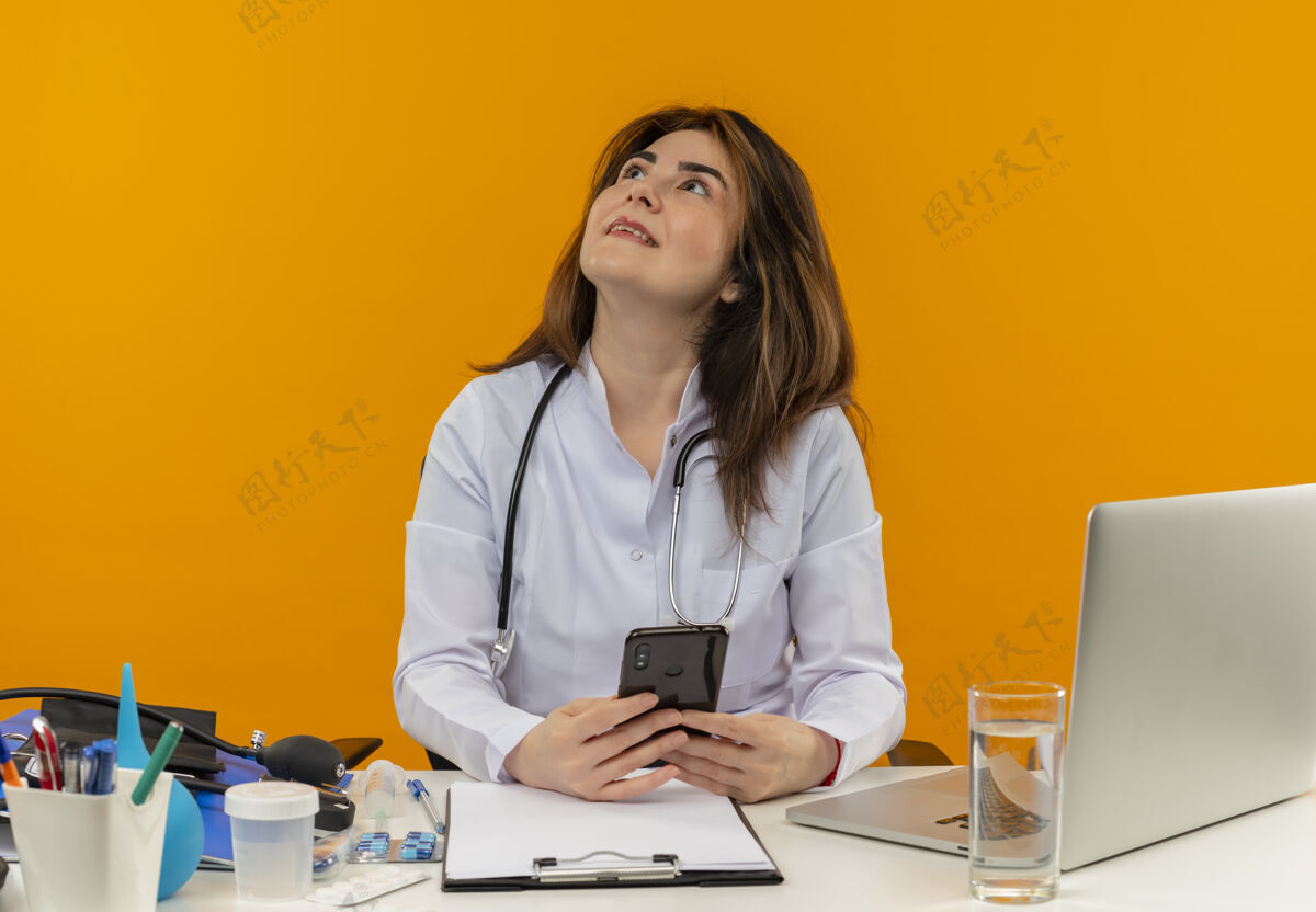拿着微笑的中年女医生穿着医用长袍和听诊器坐在办公桌旁 拿着医疗工具剪贴板和手提电脑 拿着手机孤立地抬头看笔记本电脑工具医生
