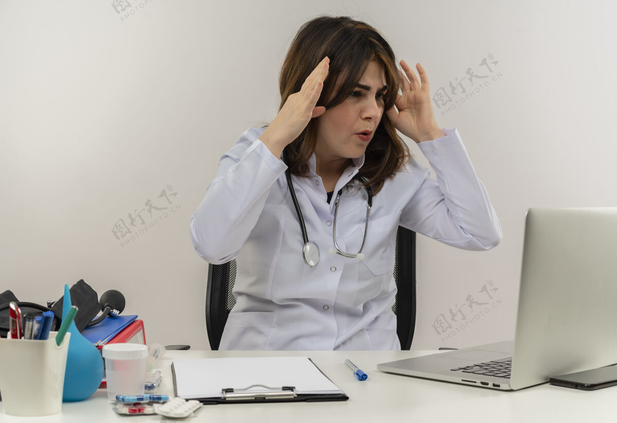 女士惊讶的中年女医生穿着医用长袍和听诊器坐在办公桌前拿着医用工具剪贴板看着笔记本电脑摸着头隔离女性桌子人