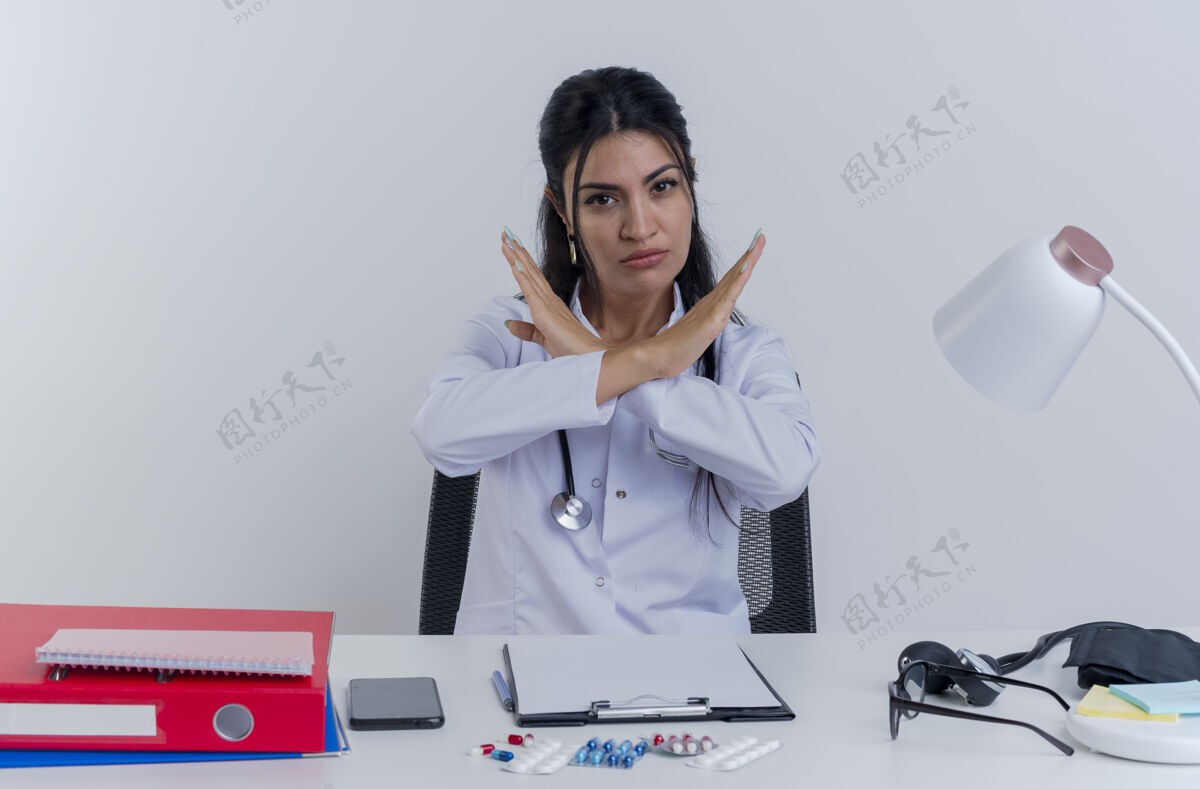医生严格的年轻女医生穿着医用长袍和听诊器坐在办公桌前 带着医疗工具 看和做没有孤立的手势医疗女士成人