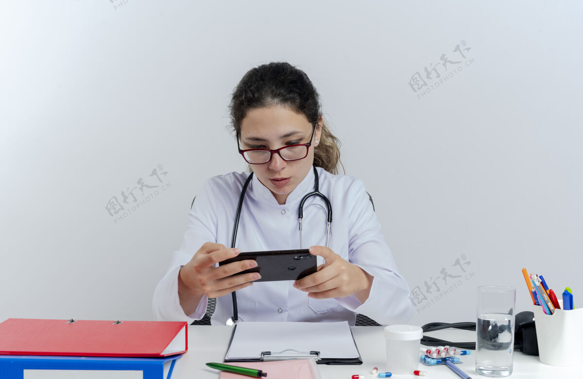 医疗年轻的女医生穿着医用长袍 戴着听诊器和眼镜坐在办公桌旁 用手机隔离医疗工具医生桌子工具