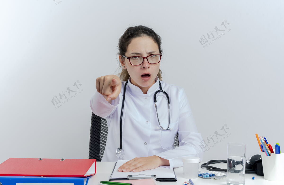 工具皱着眉头的年轻女医生穿着医用长袍 戴着听诊器和眼镜坐在办公桌旁 手里拿着医疗工具 孤立地看着和指指点点年轻人皱眉坐着的