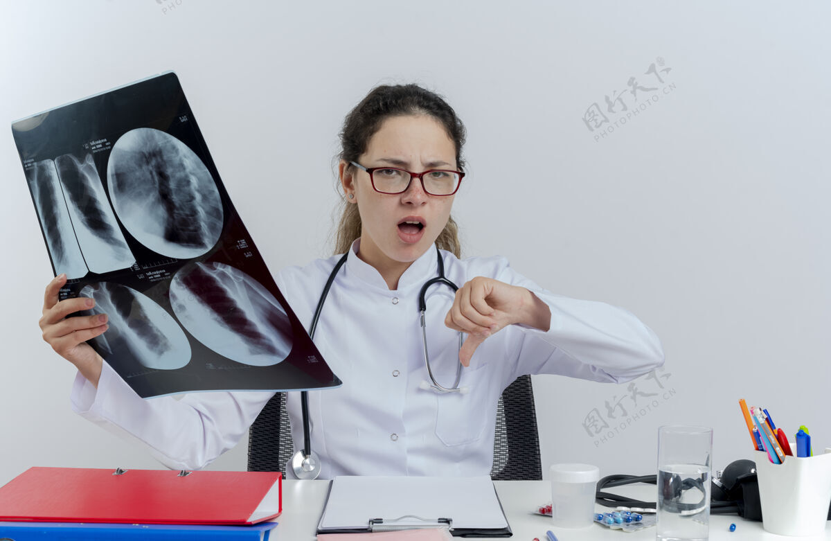 长袍年轻的女医生穿着医用长袍 戴着听诊器 戴着眼镜 坐在办公桌旁 手里拿着医疗工具 手里拿着x光片 大拇指朝下人拍摄按住