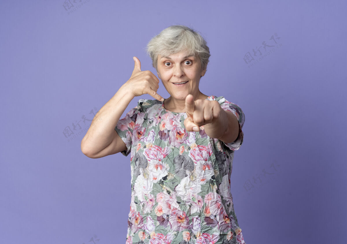 老人快乐的老妇人手势叫我的标志和点隔离在紫色的墙上手势快乐点