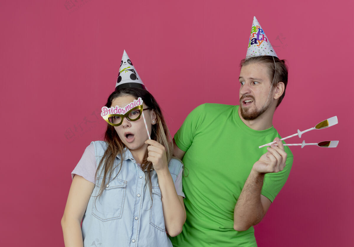 香槟不高兴的年轻人戴着派对帽拿着假香槟酒杯在棍子上看着惊讶的年轻女孩拿着眼罩在棍子上隔离在粉红色的墙上假的派对生日