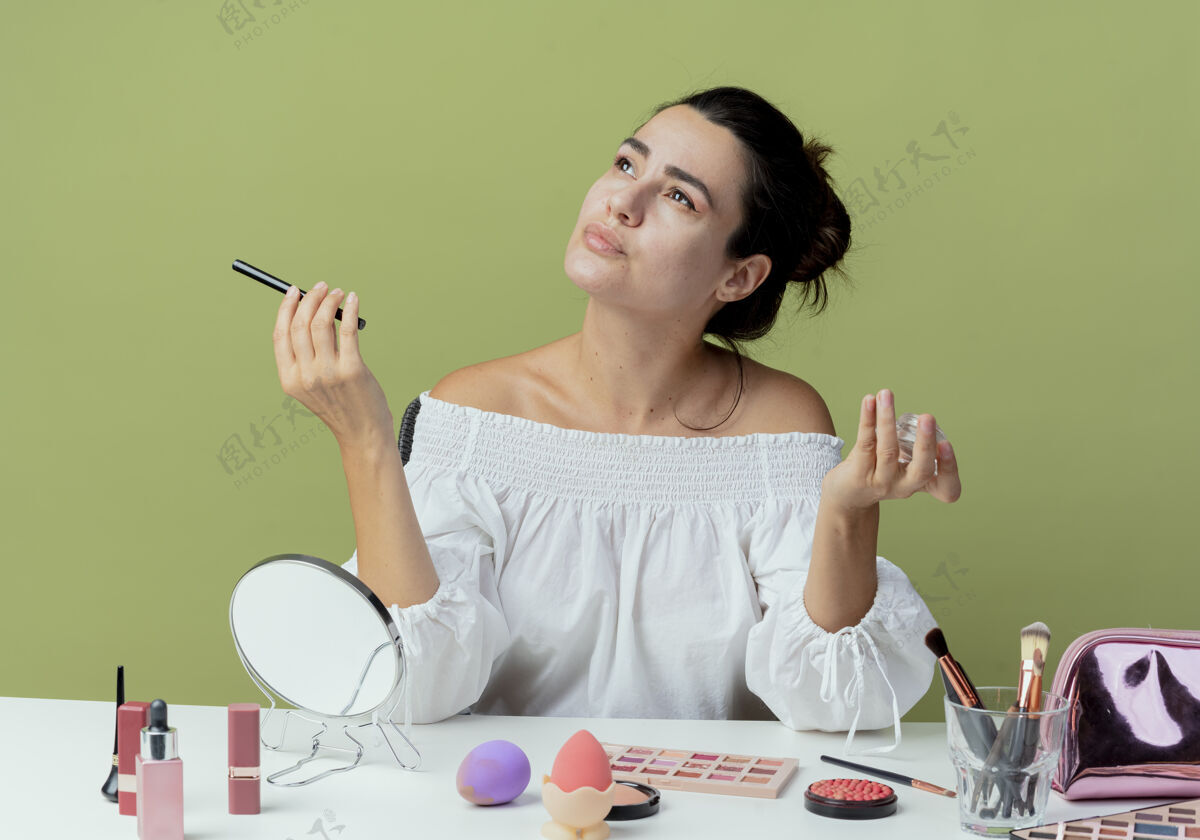 桌子自信美丽的女孩坐在桌旁 用化妆工具夹着眼影和眼线 抬头看绿色墙上的隔绝女人拿工具