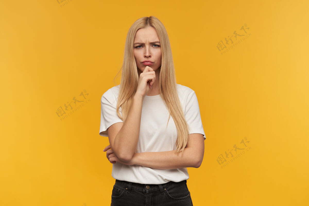 人愁眉苦脸的金发女人穿着白色t恤和黑色牛仔裤人和情感的概念看着镜头沉思 橙色背景下孤立休闲年轻成人