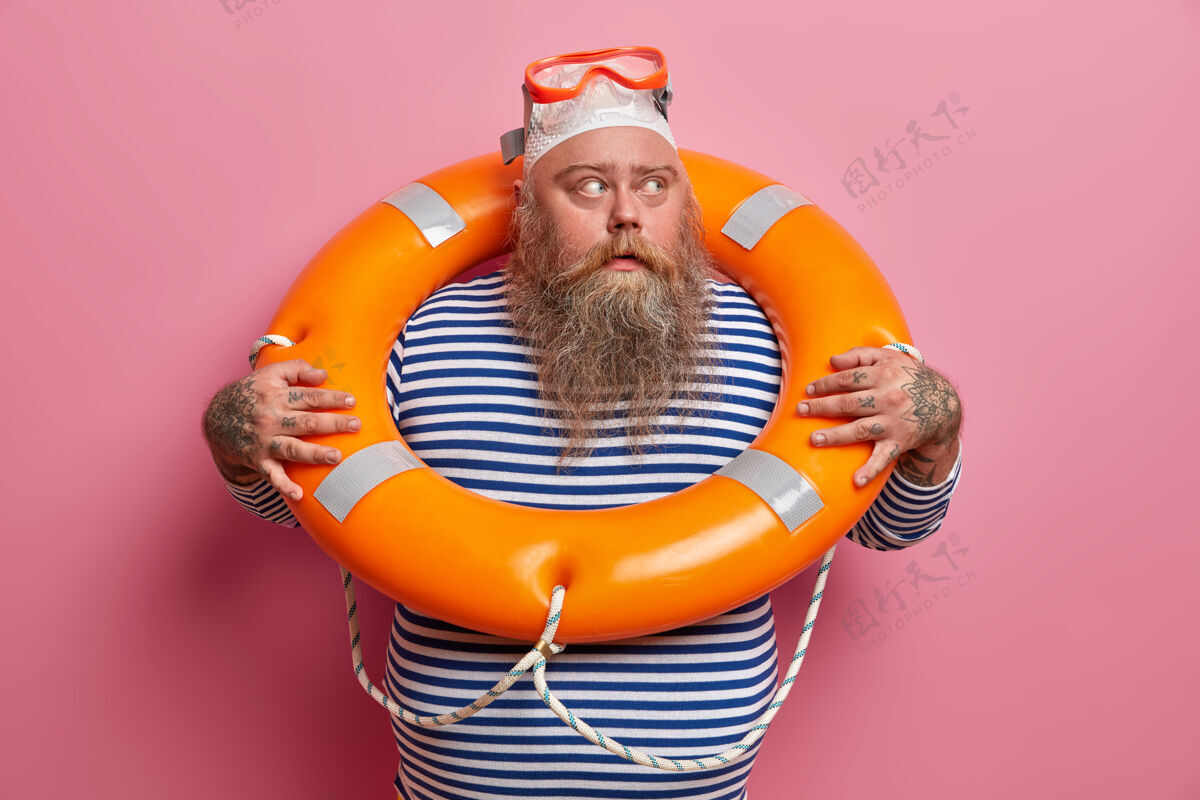 穿着戴泳帽和护目镜的大胡子男人 目光移开 穿着条纹水手背心 积极度过暑假 对着粉色墙壁摆姿势救生员值班安全海滩休息姿势室内胡须