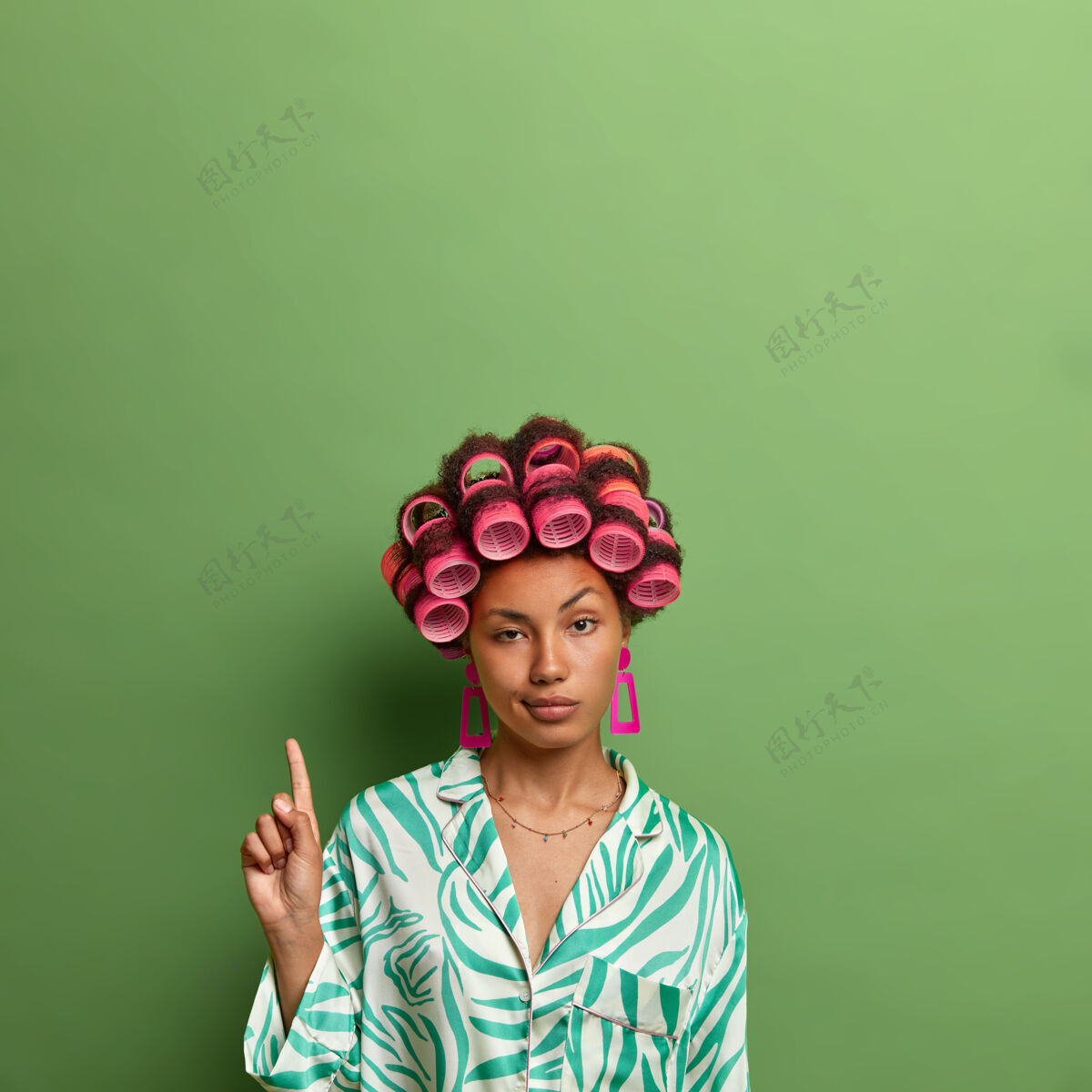女人无动于衷的黑皮肤女人的垂直镜头戴着卷发器 并在拷贝空间上标明 在绿墙的衬托下为你的宣传片显示空白 关心外表 为约会做准备站立手指姿势
