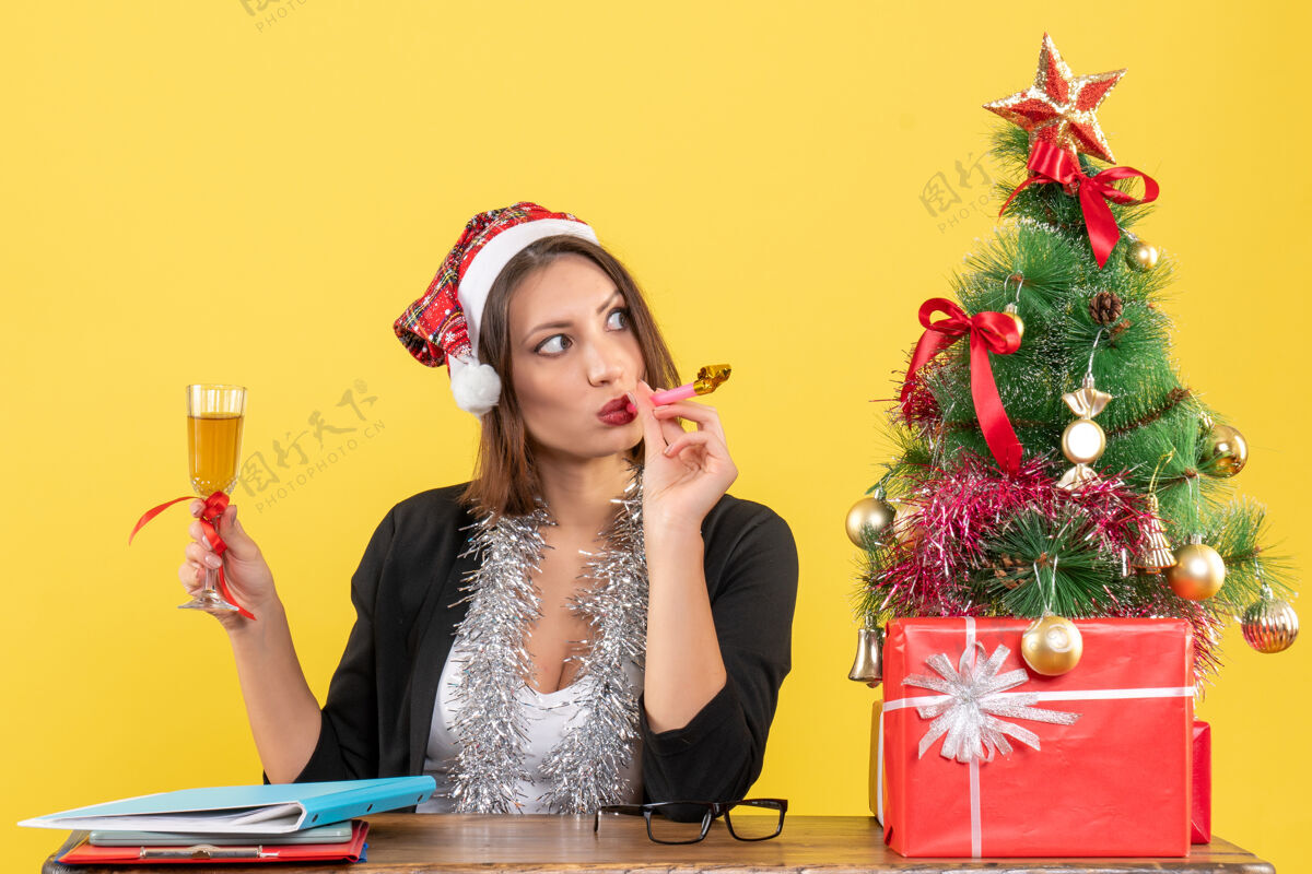 头发体贴迷人的女士穿着西装 戴着圣诞老人的帽子和新年装饰品 在黄色的办公室里举杯肖像圣诞老人虽然