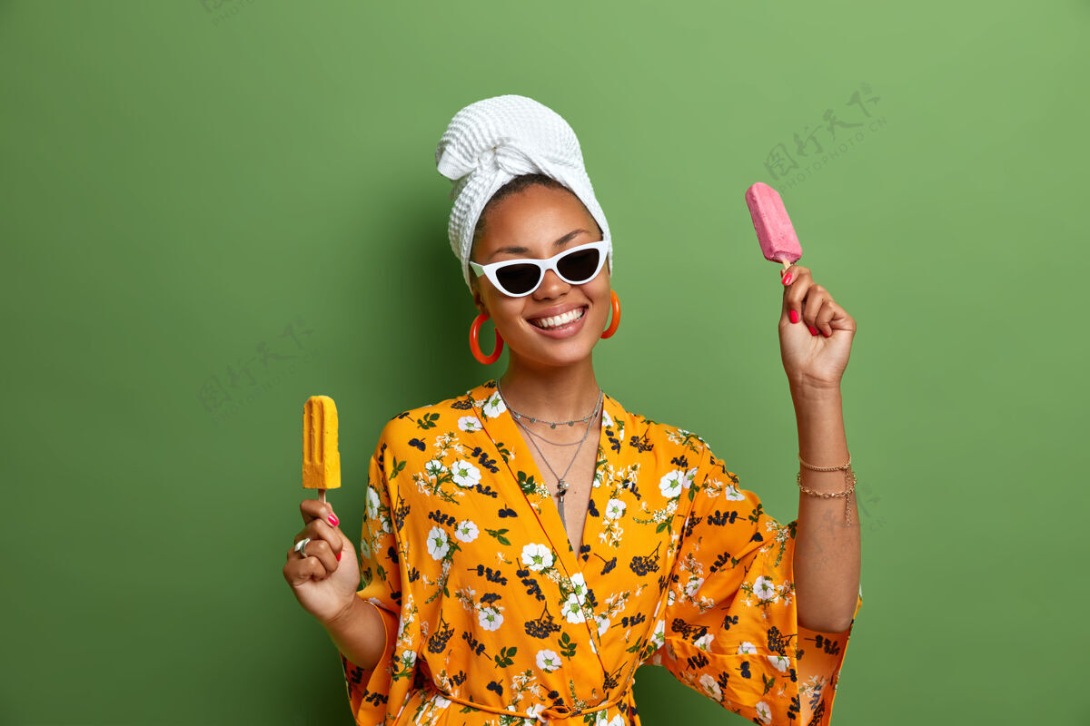 冰棒无忧无虑的积极的深色皮肤的女人拿着美味的冰淇淋 棒子上的冰棒 在夏天玩得很开心 戴着时髦的太阳镜 穿着黄色的长袍 头上裹着毛巾 喜欢吃甜食女人冰箱积极