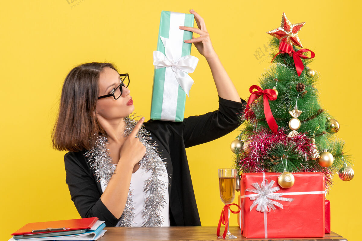 肖像一位身着西装 面带微笑的商务女士 戴着眼镜 指着她的礼物 坐在一张桌子旁 桌子上放着一棵圣诞树快乐性感树
