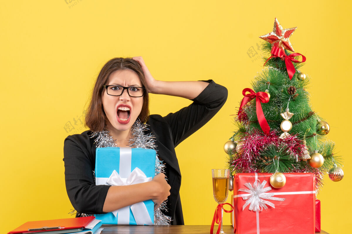成人情绪混乱的商务女士穿着西装 戴着眼镜 手里拿着礼物 坐在一张桌子旁 桌上放着一棵圣诞树漂亮头发坐着